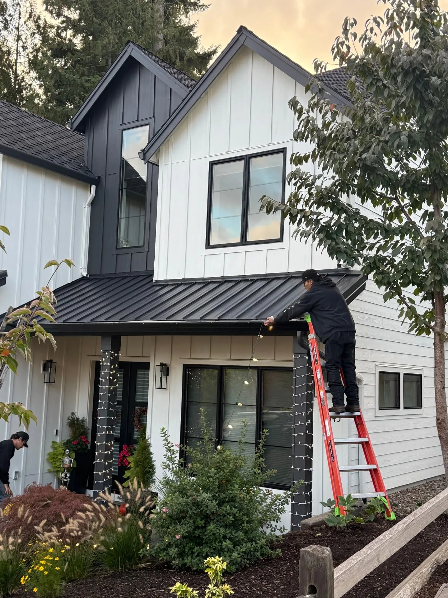North Pole Lighting Co. installer on a ladder clipping C9 bulbs along the porch roofline of a modern farmhouse while a second crew member works below