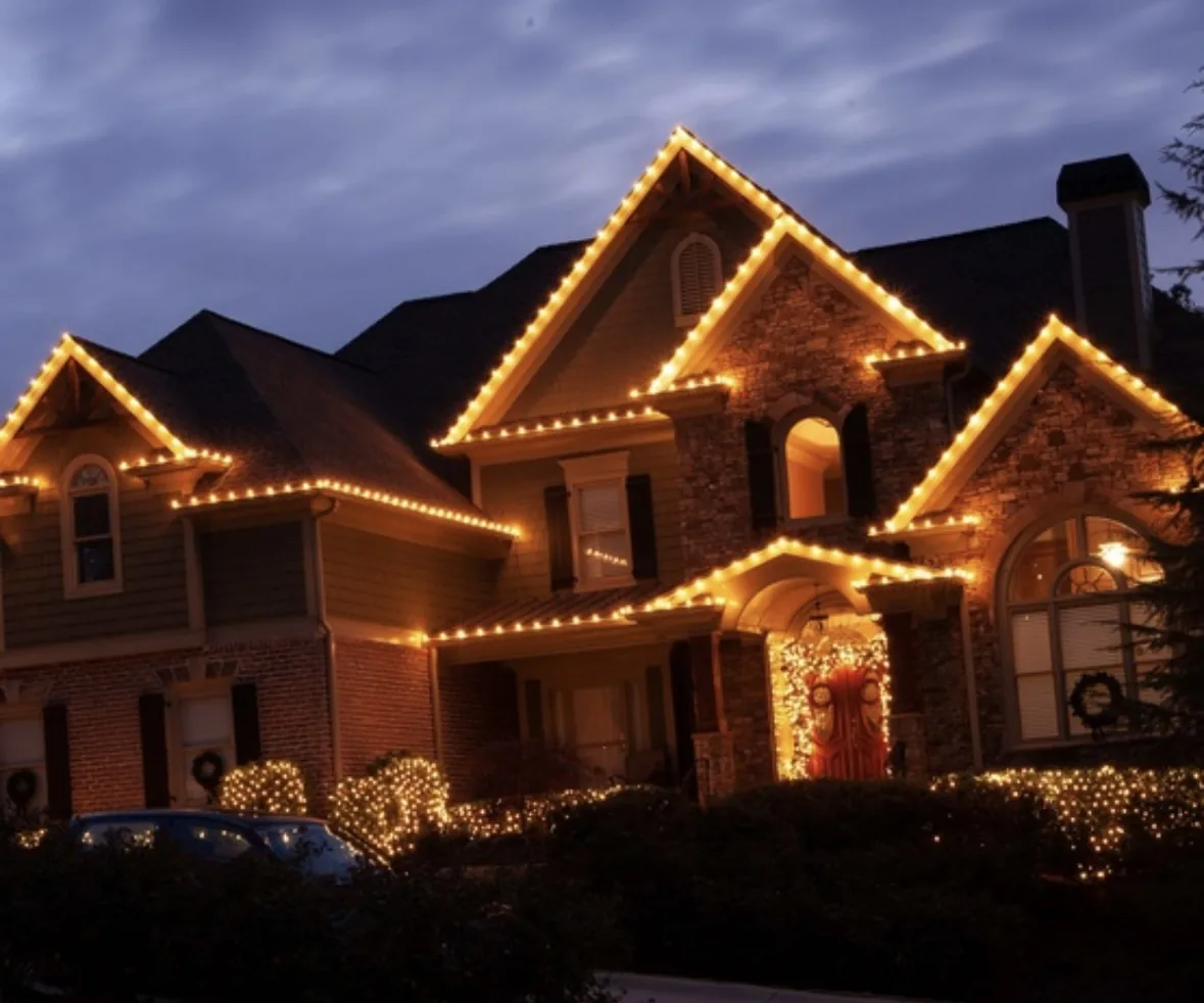 Stone and shingle home with warm white roofline lighting, lit wreaths, and net-lit shrubs at twilight
