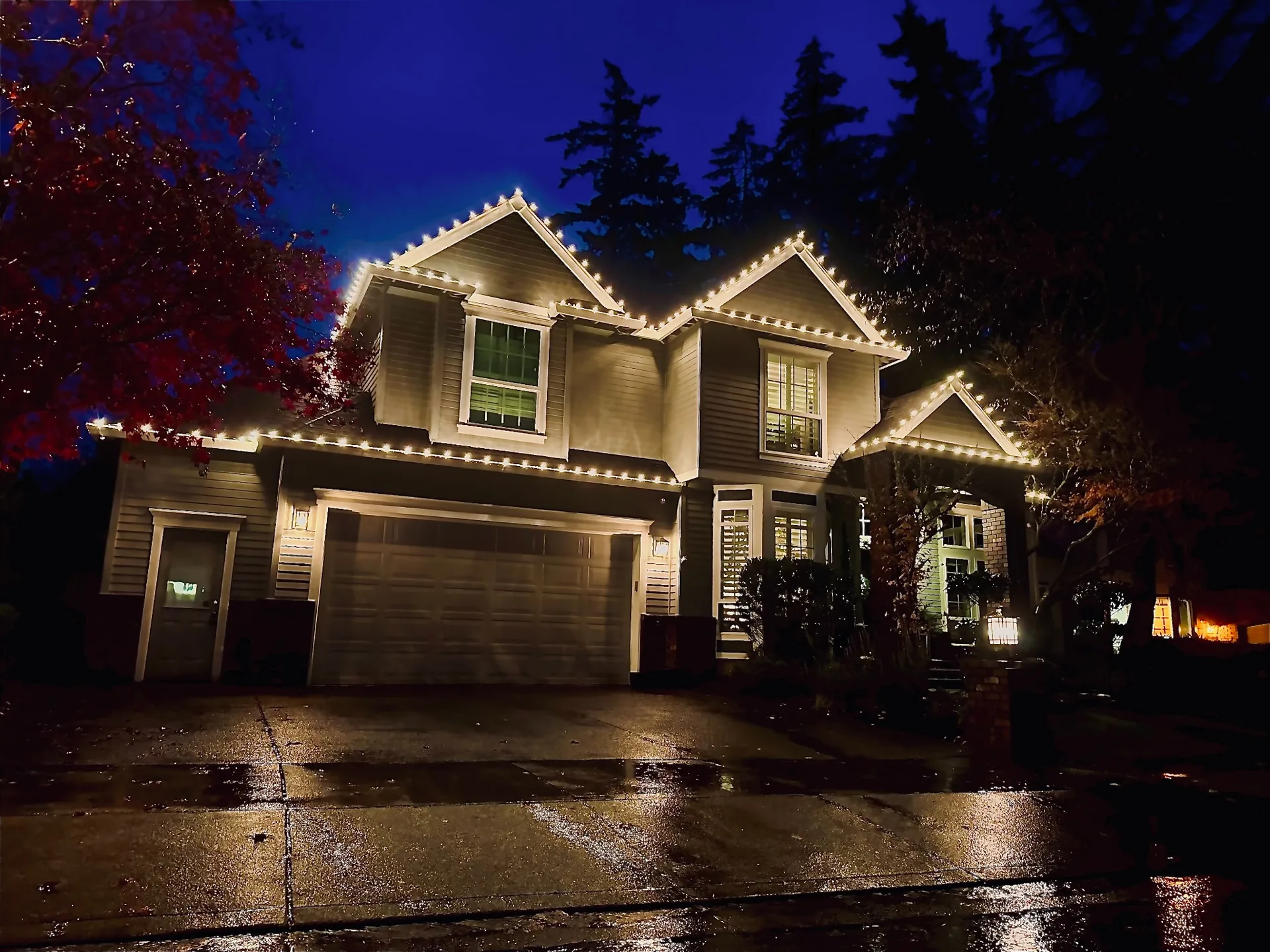 Warm white C9 holiday lights outlining every roofline and gable of a two-story Portland-area home on a wet winter evening